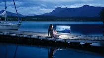 A woman sits on a dock by the water and looks at a screen. A woman sits on a dock by the water and looks at a screen.
