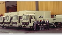 Photograph from 1986 showing five BASF delivery lorries parked outside an industrial warehouse, with five employees standing in front of the vehicles. BASF branding is visible on the trucks and warehouse loading area. Photograph from 1986 showing five BASF delivery lorries parked outside an industrial warehouse, with five employees standing in front of the vehicles. BASF branding is visible on the trucks and warehouse loading area.