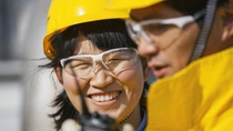 Close-up of two workers wearing yellow safety helmets and protective glasses, collaborating outdoors; one smiles while looking at a component or tool, the other focuses on the task in the foreground. Close-up of two workers wearing yellow safety helmets and protective glasses, collaborating outdoors; one smiles while looking at a component or tool, the other focuses on the task in the foreground.
