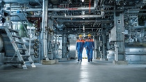 Zwei Personen in Schutzkleidung und Helmen gehen durch eine Industrieanlage mit Rohrleitungen und Anlagen – chemische Produktion und Arbeitssicherheit. Zwei Personen in Schutzkleidung und Helmen gehen durch eine Industrieanlage mit Rohrleitungen und Anlagen – chemische Produktion und Arbeitssicherheit.