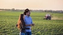 A man and woman standing on a field with a tractor in the background.