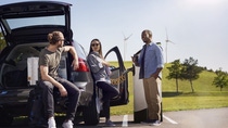 A group of three people, two men and one women, is chatting casually and drinking coffee, while their EV is charging at a charging station. In the backdrop, one can see green hills and some windmills.  A group of three people, two men and one women, is chatting casually and drinking coffee, while their EV is charging at a charging station. In the backdrop, one can see green hills and some windmills.