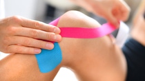 Kinesiology taping treatment with blue and pink tape on athlete patient injured knee. Woman hands apply kinesio treatment after sports muscle injury. Kinesiology taping treatment with blue and pink tape on athlete patient injured knee. Woman hands apply kinesio treatment after sports muscle injury.