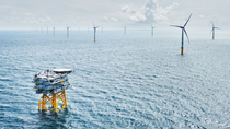 A wide view of a blue ocean with waves taken from above, showing two rows of wind turbines stretching into the distance and an offshore platform with helicopter landing pad in the foreground.  A wide view of a blue ocean with waves taken from above, showing two rows of wind turbines stretching into the distance and an offshore platform with helicopter landing pad in the foreground.