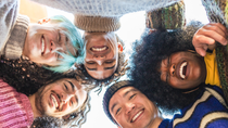 A diverse group of people in casual clothing lean together in a circle, smiling and looking down at the camera, representing connection, inclusion, and team spirit. A diverse group of people in casual clothing lean together in a circle, smiling and looking down at the camera, representing connection, inclusion, and team spirit.