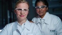 Two female scientists wearing white lab coats with the BASF logo and safety goggles stand in the laboratory and look into the camera. Two female scientists wearing white lab coats with the BASF logo and safety goggles stand in the laboratory and look into the camera.