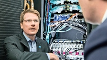 Ein Mann sitzt vor Computerkabeln und spricht Jemanden abseits der Kamera an. Ein Mann sitzt vor Computerkabeln und spricht Jemanden abseits der Kamera an.