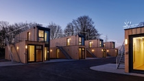A small village of wooden-cladded tiny houses with large windows made from upcycled shipping containers stacked one on top of another. It is dusk and warm-colored lights are illuminating the interiors. (Photo: Containerwerk)  A small village of wooden-cladded tiny houses with large windows made from upcycled shipping containers stacked one on top of another. It is dusk and warm-colored lights are illuminating the interiors. (Photo: Containerwerk)
