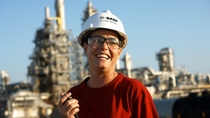 A BASF employee wearing a safety helmet and protective glasses stands at an industrial site, smiling and holding a walkie-talkie, with production facilities in the background. A BASF employee wearing a safety helmet and protective glasses stands at an industrial site, smiling and holding a walkie-talkie, with production facilities in the background.