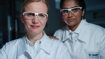 Two female scientists wearing white lab coats with the BASF logo and safety goggles stand in the laboratory and look into the camera. Two female scientists wearing white lab coats with the BASF logo and safety goggles stand in the laboratory and look into the camera.