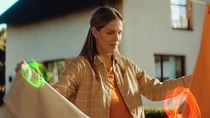 Une femme se tient debout à l’extérieur, décrochant une nappe d’un filà linge devant une maison avec de la végétation en arrière-plan. Des tourbillons lumineux flottent près de ses mains tenant le linge. Une femme se tient debout à l’extérieur, décrochant une nappe d’un filà linge devant une maison avec de la végétation en arrière-plan. Des tourbillons lumineux flottent près de ses mains tenant le linge.