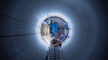 Blick von unten in einen großen, zylindrischen Behälter, in dem mehrere Arbeiter mit Sicherheitsausrüstung an der Innenwand arbeiten; eine Leiter führt nach oben. Blick von unten in einen großen, zylindrischen Behälter, in dem mehrere Arbeiter mit Sicherheitsausrüstung an der Innenwand arbeiten; eine Leiter führt nach oben.