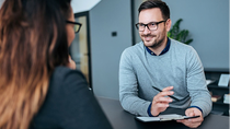 Ein freundlicher Berater im grauen Pullover führt ein persönliches Gespräch mit einer Kundin in einem modernen Büro, während er Notizen auf einem Klemmbrett macht. Ein freundlicher Berater im grauen Pullover führt ein persönliches Gespräch mit einer Kundin in einem modernen Büro, während er Notizen auf einem Klemmbrett macht.