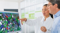 Two scientists, one man and one woman, stand together looking at a 3D scientific diagram on a computer screen. Two scientists, one man and one woman, stand together looking at a 3D scientific diagram on a computer screen.