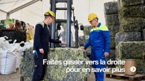 Deux hommes portant des casques jaunes et une salopette de travail se tiennent à côté d’un grand bloc pressé de déchets textiles.  Deux hommes portant des casques jaunes et une salopette de travail se tiennent à côté d’un grand bloc pressé de déchets textiles.