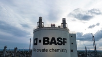 Wide view of an industrial BASF facility with a large storage tank labeled “BASF – We create chemistry,” where several workers in safety helmets stand together on the top platform under a cloudy sky. Wide view of an industrial BASF facility with a large storage tank labeled “BASF – We create chemistry,” where several workers in safety helmets stand together on the top platform under a cloudy sky.