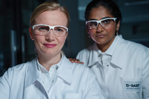 Two female scientists in white lab coats with the BASF logo and protective goggles stand in the laboratory and look into the camera. Two female scientists in white lab coats with the BASF logo and protective goggles stand in the laboratory and look into the camera.