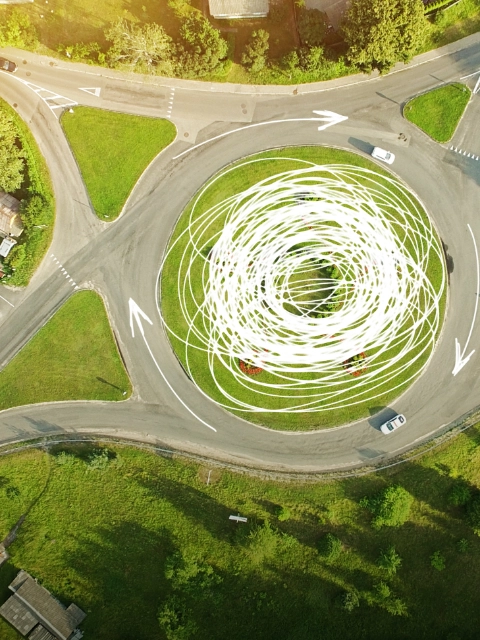 Aerial view of a grassy roundabout with four exits, surrounded by trees and green fields - scientific equations and sketches highlighting the flow of traffic are overlaid on the picture.