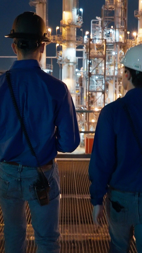 Two men with satety helmets are walking side by side in a lit production facility Two men with satety helmets are walking side by side in a lit production facility
