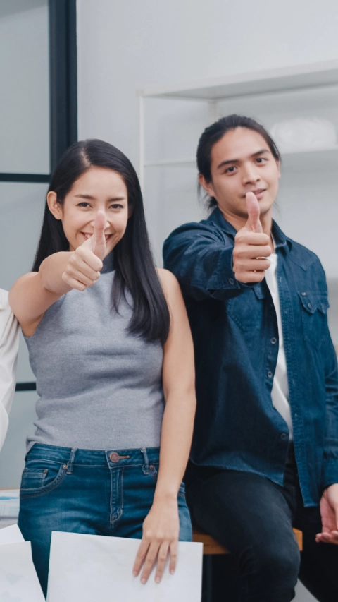 Group of Asia young creative people in smart casual wear smiling and thumbs up in creative office workplace. Diverse Asian male and female stand together at startup. Coworker teamwork concept.