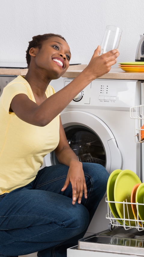 Happy African Woman Looking At Clean Glass Crouching Near Dish Washer Happy African Woman Looking At Clean Glass Crouching Near Dish Washer