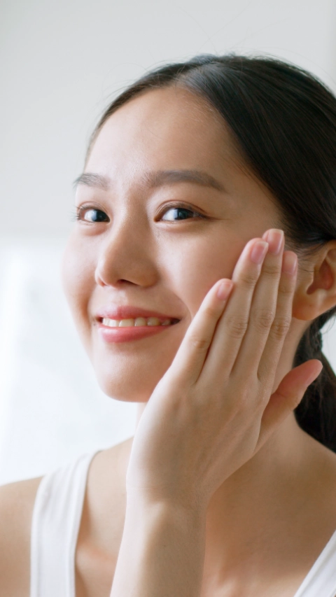 Young Asian woman touching healthy facial beauty skin look at mirror